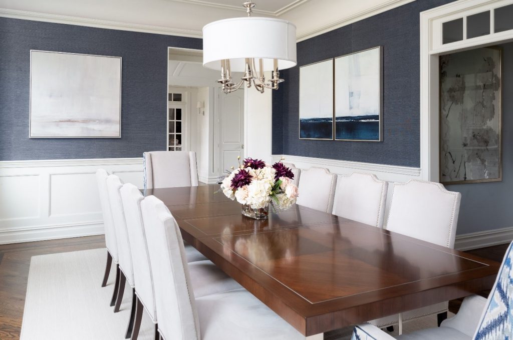 Dining room design, NJ Interior Designer. Neutral artwork, artwork with dark blue. Low hung light fixture, long rectangular dining table, host chairs, side chairs, blue grasscloth wallpaper, white wall paneling and floral decor on table. 