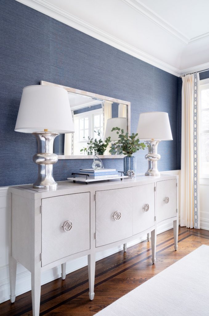 Dining room design, NJ Interior Designer. Blue grasscloth wallpaper, matching lamps, off white buffet with reading materials and a small plant, hung above the buffet a rectangular mirror. 