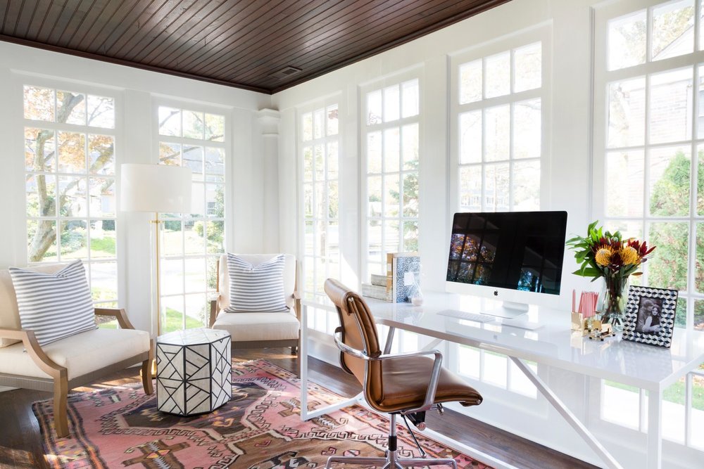 Home office with natural light, seating area, white desk, brown chair, multi-colored rug and large windows.