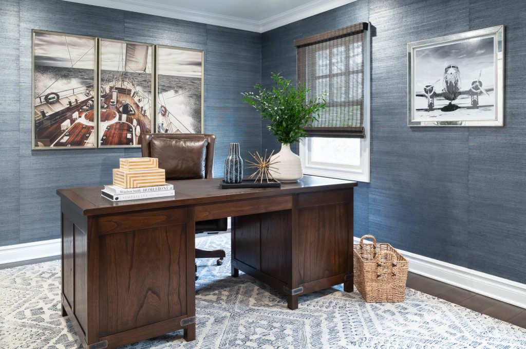 home office with brown desk, artwork on walls, blue wallpaper, plant on desk, decor on desk, woven basket on floor.