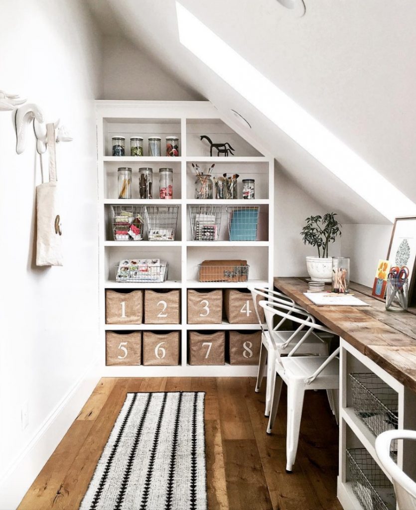 Office with built in book shelf with decor and bins. Long wooden desk, white chairs, hardwood floors and patterned rug. 