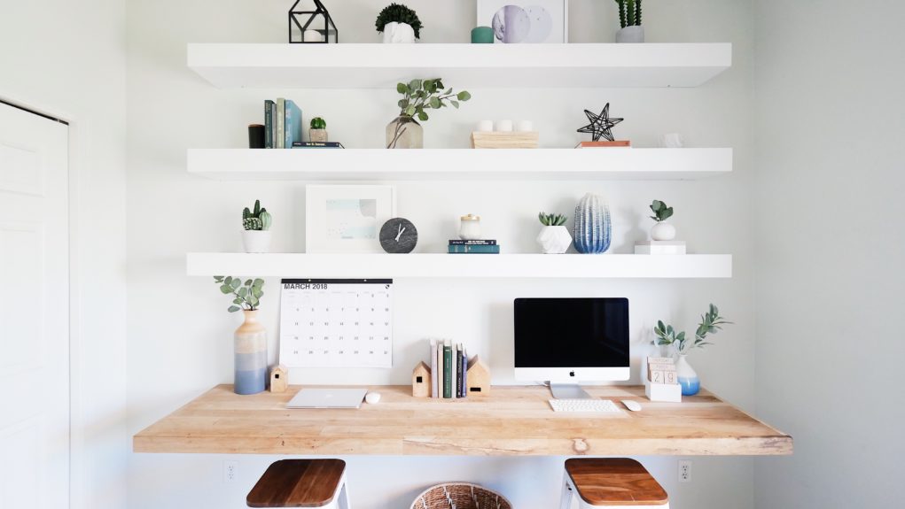 White floating shelves with plants and decor. Wood desk with office supplies.